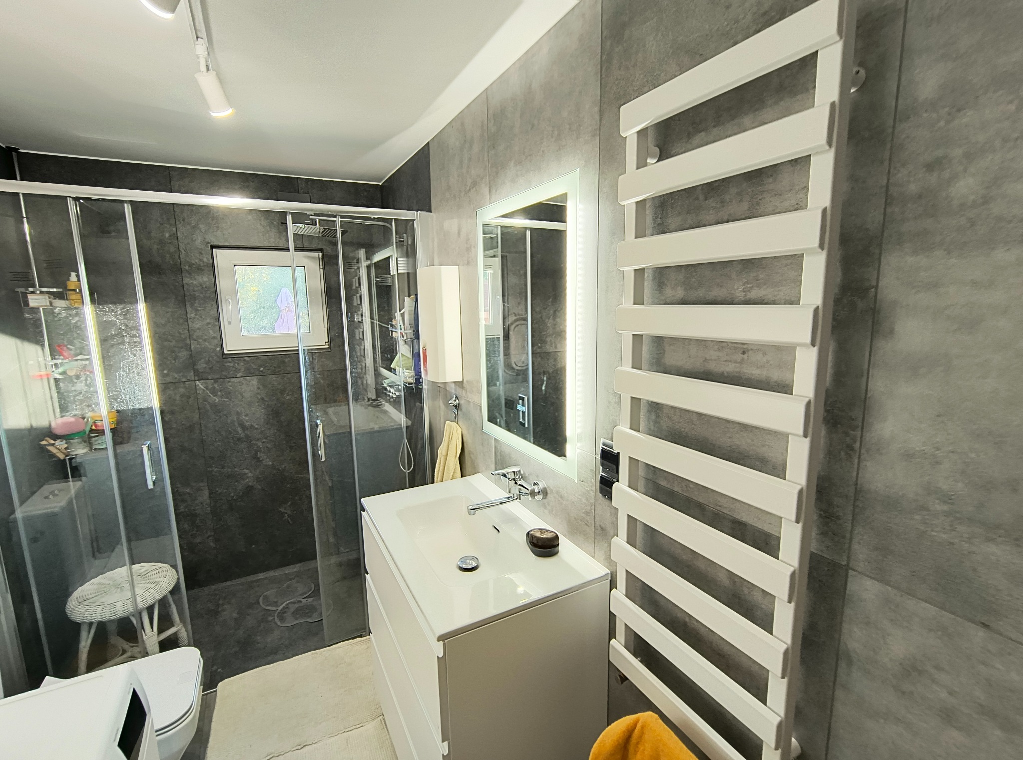 Nowoczesna łazienka z szarą okładziną, kabiną prysznicową ze szklanymi drzwiami, białą szafką z umywalką, podświetlanym lustrem i grzejnikiem drabinkowym.