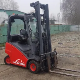 Czerwono-szary wózek widłowy Linde na betonowym placu z widocznym budynkiem i ogrodzeniem w tle, w tle unoszący się dym.