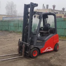 Czerwono-szary wózek widłowy Linde na betonowym placu, w tle zielone ogrodzenie i budynki spowite mgłą.