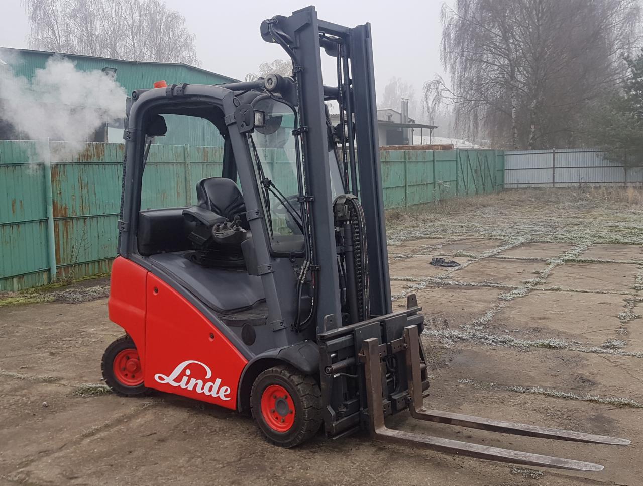 Czerwono-szary wózek widłowy Linde na betonowym placu z widocznym budynkiem i ogrodzeniem w tle, w tle unoszący się dym.