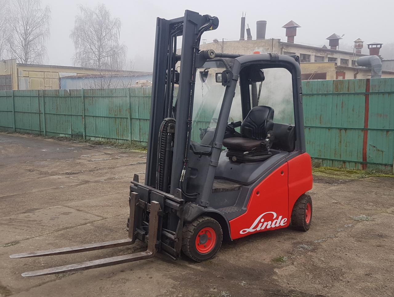 Czerwono-szary wózek widłowy Linde na betonowym placu, w tle zielone ogrodzenie i budynki spowite mgłą.