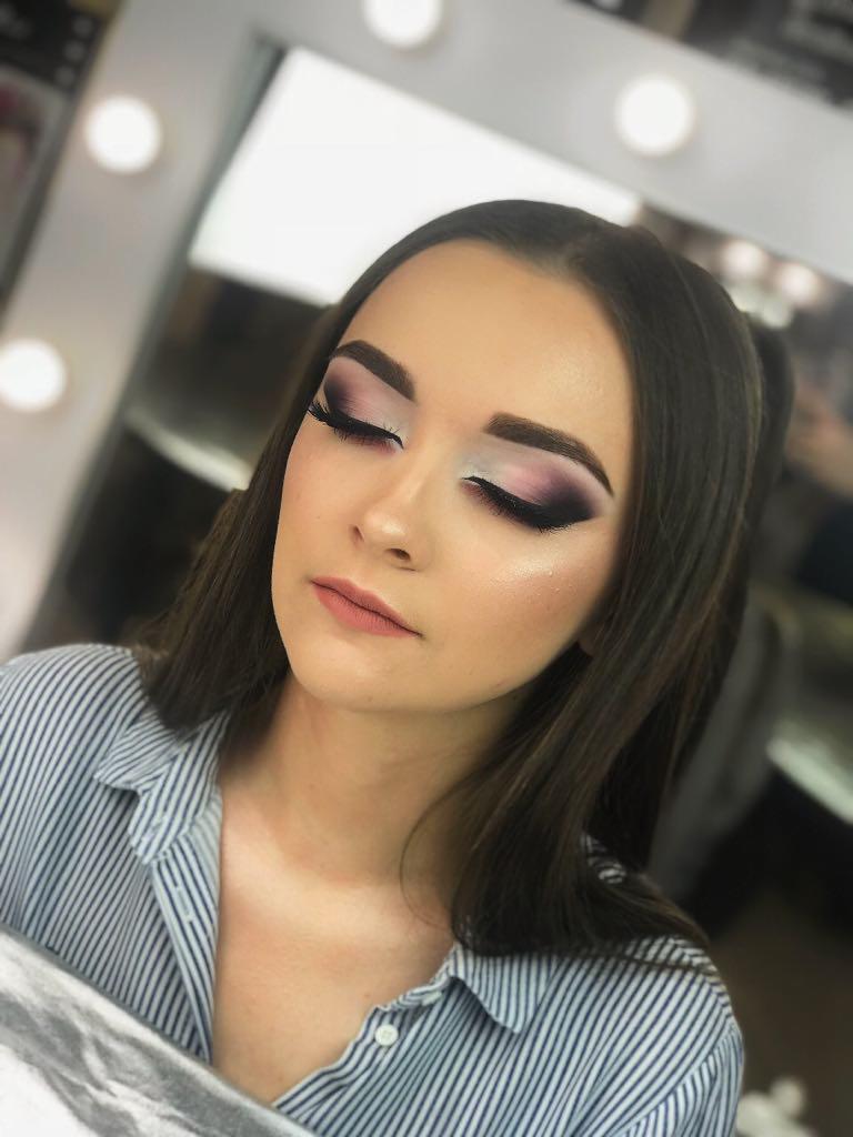 Portret kobiety z zamkniętymi oczami, prezentujący delikatny, fioletowo-biały makijaż oczu z długimi, sztucznymi rzęsami i subtelnym konturowaniem twarzy, w tle oświetlone lustro.