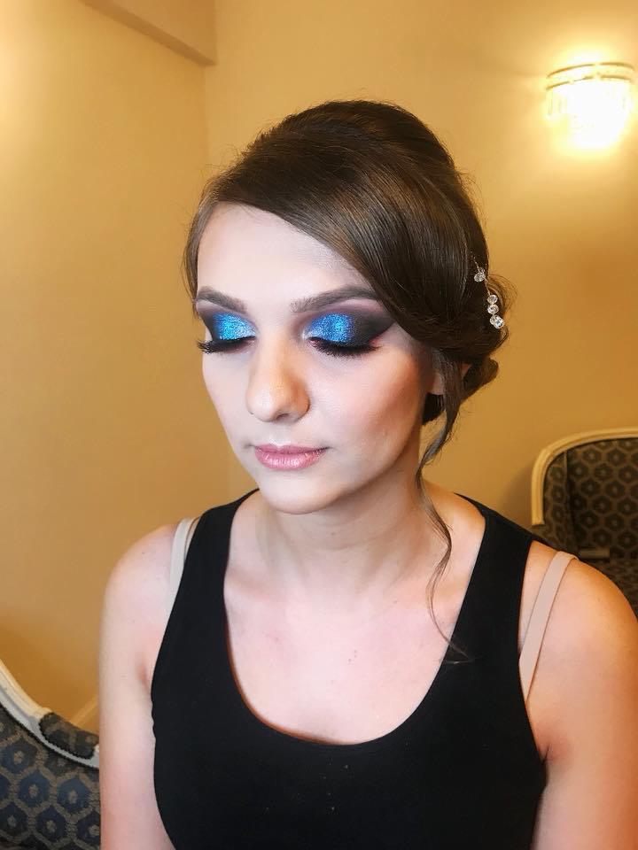 Portret kobiety z wieczorowym upięciem włosów i intensywnym, błyszczącym niebieskim cieniem na powiekach oraz sztucznymi rzęsami. W tle widoczny fragment krzesła w stylu vintage.