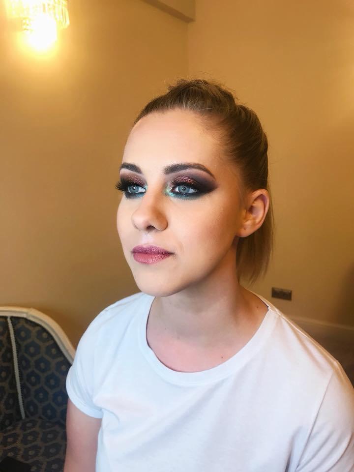 Portret kobiety z intensywnym, brokatowym makijażem oczu w odcieniach fioletu, zieleni i brązu, z wyraźnie podkreślonymi rzęsami i delikatnym różem na ustach, spięte włosy, biała koszulka