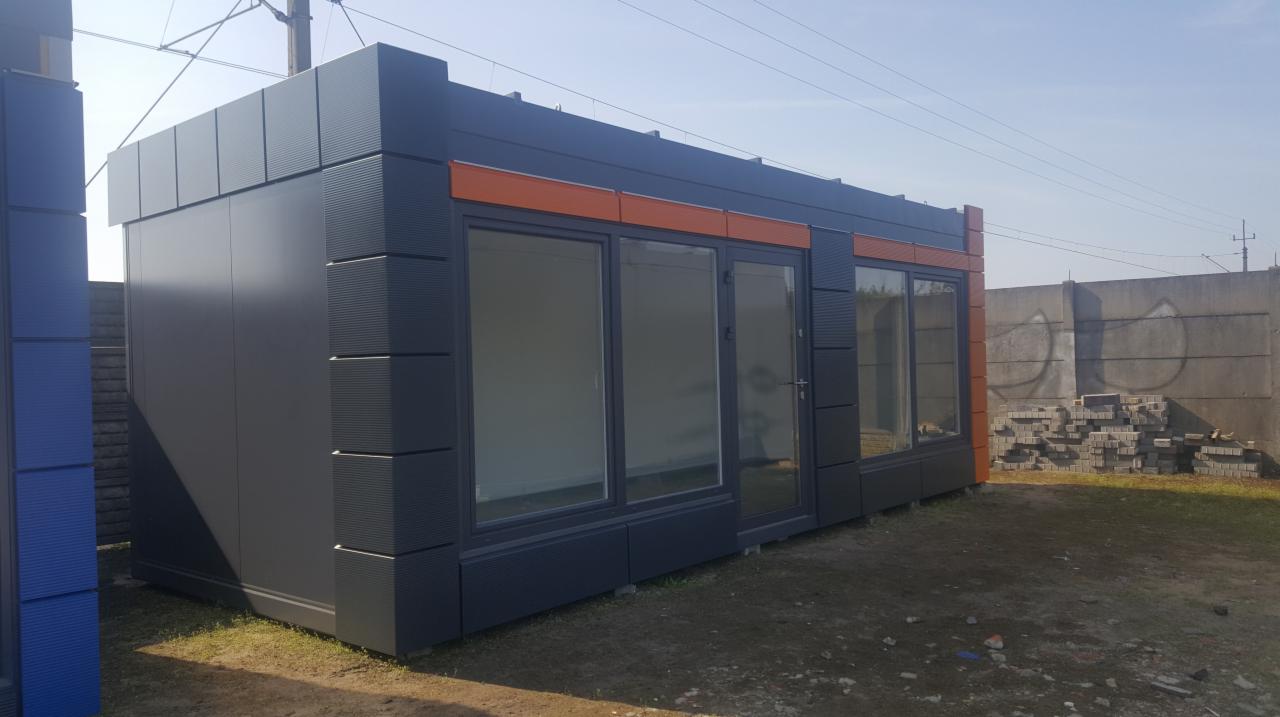 Nowoczesny pawilon handlowy z szarej blachy trapezowej z pomarańczowymi akcentami, widok z zewnątrz, na ziemi leżą kostki brukowe.