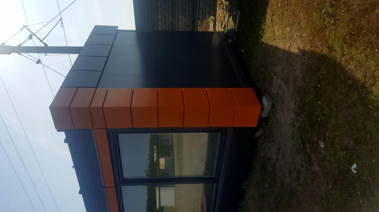 Nowoczesny pawilon handlowy z ciemnoszarą elewacją i pomarańczowymi elementami dekoracyjnymi, widoczny z zewnątrz na tle trawy i słupa energetycznego.