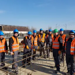 Grupa dziewięciu robotników budowlanych w kaskach (jeden zielony, reszta niebieskie) i pomarańczowych kamizelkach odblaskowych, stojąca na placu budowy na tle zbrojenia i budynku przemysłowego.