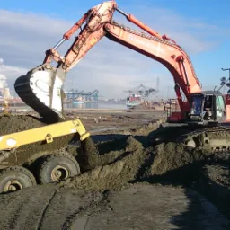 Pomarańczowa koparka Herbosch-Kiere Hitachi załadowuje urobek do żółtej wywrotki Volvo na placu budowy z dźwigami i infrastrukturą portową w tle.