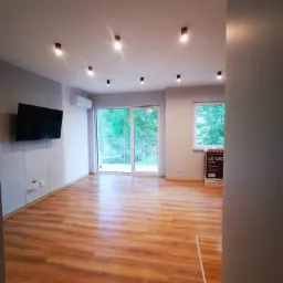 Jasne, przestronne wnętrze po remoncie w Mrągowie z drewnianą podłogą, szarą ścianą z panelami imitującymi beton, nowoczesnym oświetleniem punktowym i dużymi oknami balkonowymi z widokiem na zieleń.