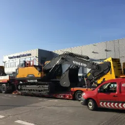 Ż&oacute;łta koparka Volvo na niskopodwoziowej platformie transportowana przez ciężar&oacute;wkę, z czerwonym vanem Witkowski Transport z boku, na tle budynku Volvo Maszyny Budowlane.