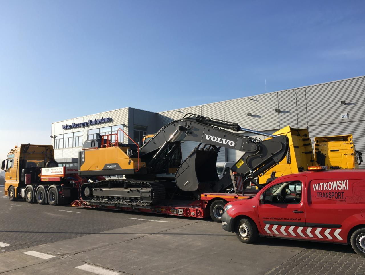 Żółta koparka Volvo na niskopodwoziowej platformie transportowana przez ciężarówkę, z czerwonym vanem Witkowski Transport z boku, na tle budynku Volvo Maszyny Budowlane.