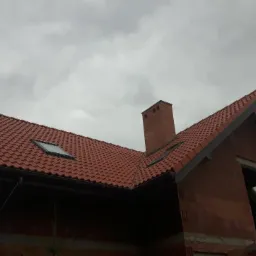 Dom w trakcie budowy z nowym, czerwonym dachem z dachówki ceramicznej i oknami dachowymi, widok z dołu na tle pochmurnego nieba.