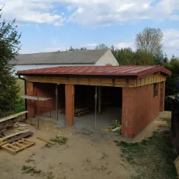 Wznoszona z czerwonej cegły ściana garażu z widocznym drewnianym szalunkiem pod dach, częściowo pokrytym blachodachówką w kolorze bordowym, na piaszczystym podłożu, z widocznymi elementami...