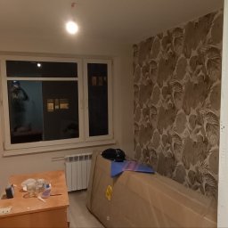 Oscar - Pokój w trakcie remontu: widoczne świeżo pomalowane ściany w odcieniach bieli, okno z parapetem, grzejnik, meble zabezpieczone folią, na stoliku szklanki i drobne narzędzia.