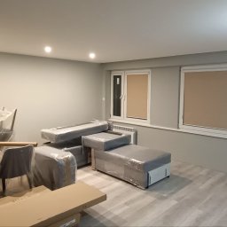 Oscar - Pokój w trakcie remontu: szare ściany, okna z roletami, meble zabezpieczone folią, widoczne przewody elektryczne na ścianie.