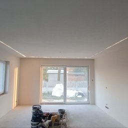 Oscar - Wnętrze pokoju po szpachlowaniu, z widocznymi śladami po gładzi na ścianach i suficie, oraz przygotowanymi narzędziami i wiadrami na podłodze. Sufit z zamontowanym oświetleniem LED wzdłuż krawędzi.