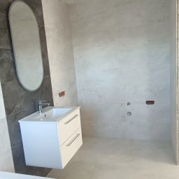 Oscar - Nowoczesna łazienka w trakcie wykańczania, widoczna biała szafka z umywalką, owalne lustro na szarej ścianie i szare płytki na ścianach oraz podłodze; brakujące elementy instalacji.