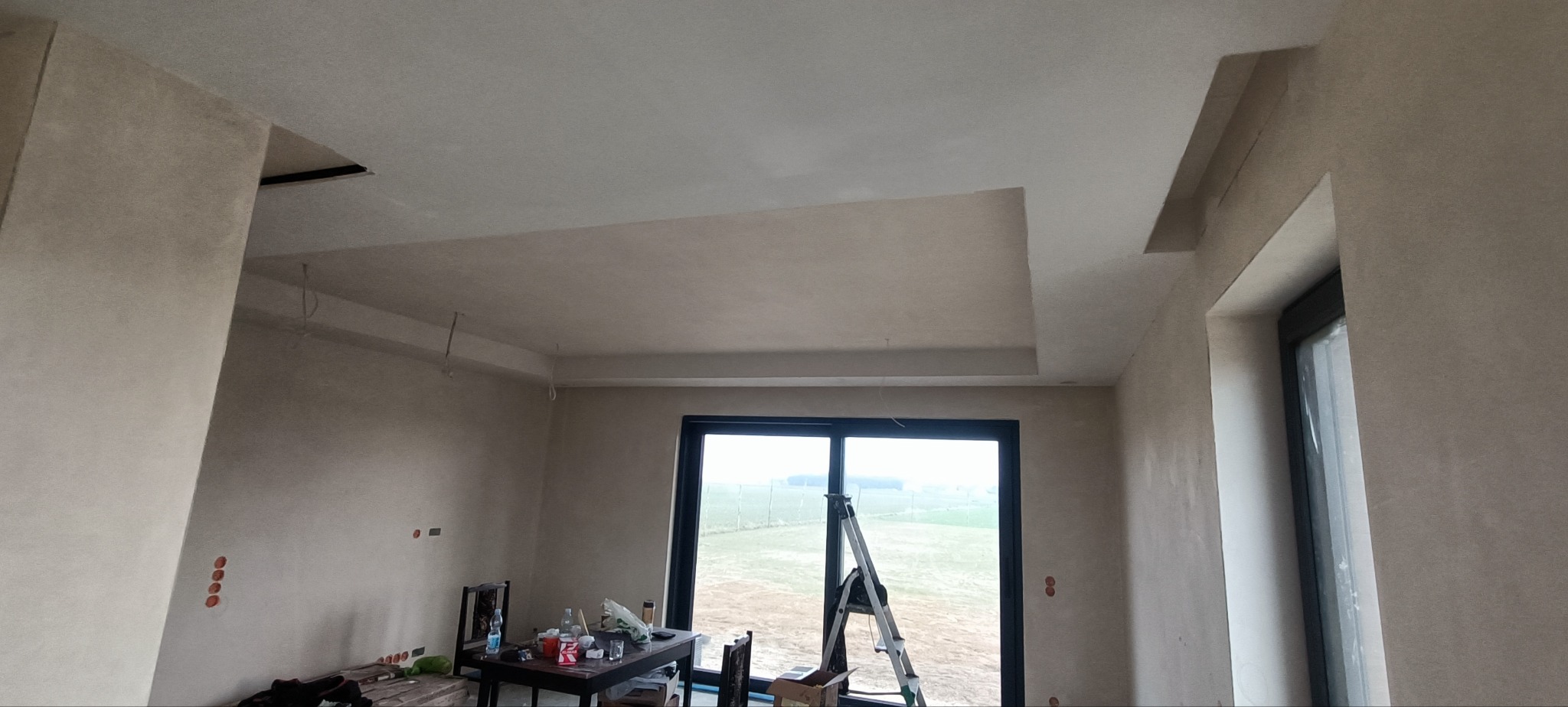 Wnętrze w trakcie prac wykończeniowych: ściany i sufit po szpachlowaniu, widoczne niedoskonałości i nierówności, przygotowane do dalszych prac, drabina oparta o okno, stół z akcesoriami budowlanymi...