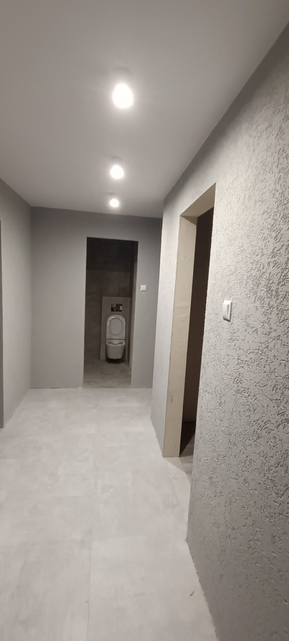 Wnętrze w odcieniach szarości, z widoczną toaletą w głębi, ściana z wyraźną fakturą i świeżo pomalowane powierzchnie.