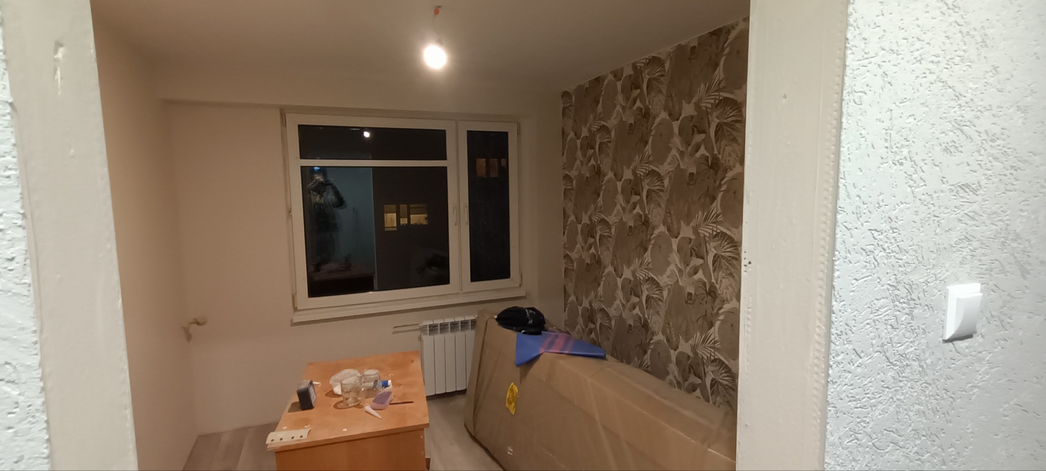 Pokój w trakcie remontu: widoczne świeżo pomalowane ściany w odcieniach bieli, okno z parapetem, grzejnik, meble zabezpieczone folią, na stoliku szklanki i drobne narzędzia.