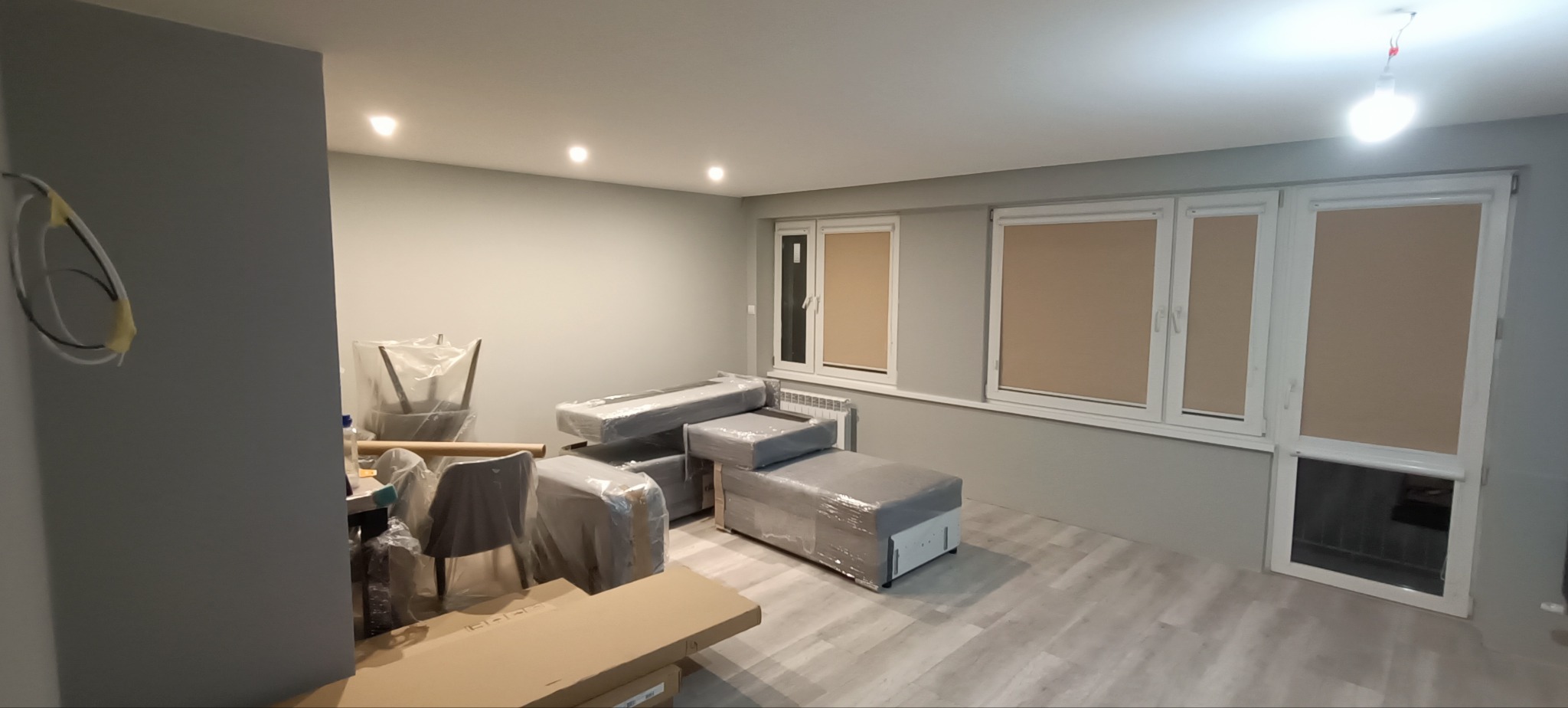 Pokój w trakcie remontu: szare ściany, okna z roletami, meble zabezpieczone folią, widoczne przewody elektryczne na ścianie.