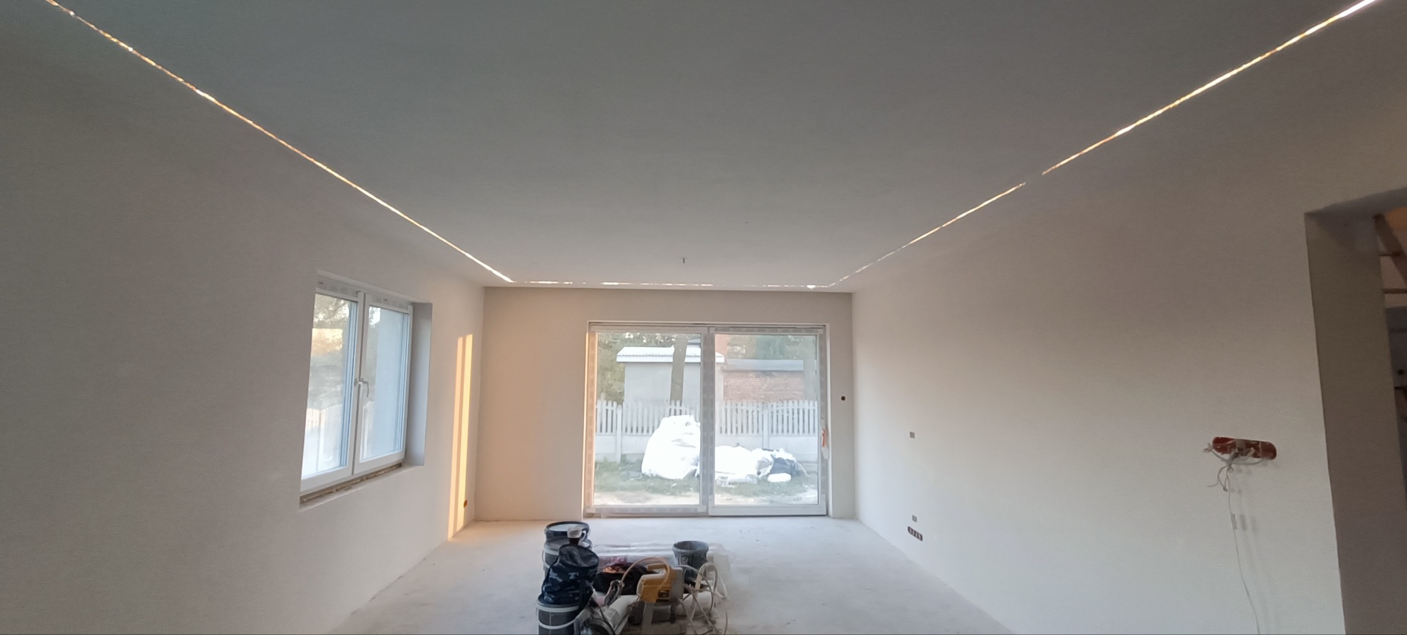 Wnętrze pokoju po szpachlowaniu, z widocznymi śladami po gładzi na ścianach i suficie, oraz przygotowanymi narzędziami i wiadrami na podłodze. Sufit z zamontowanym oświetleniem LED wzdłuż krawędzi.