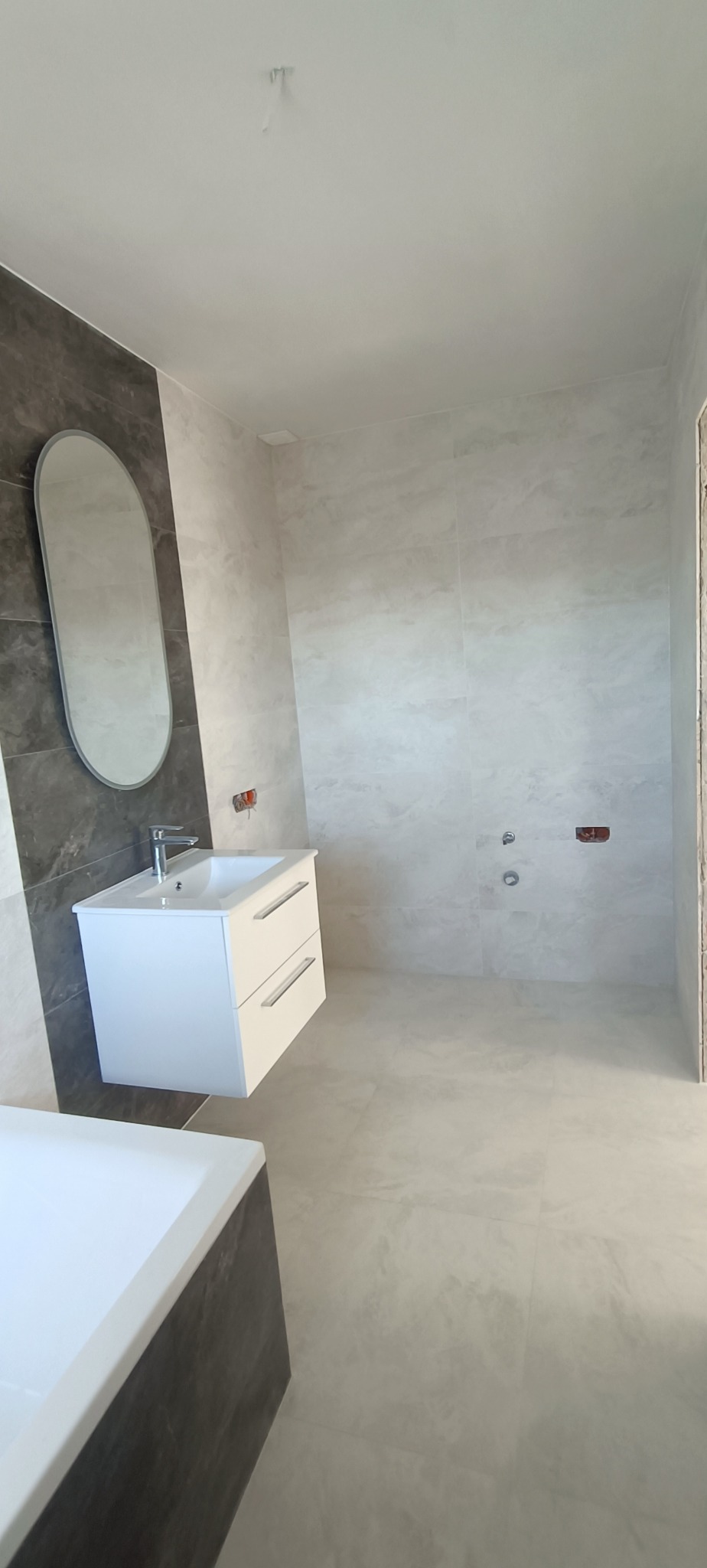 Nowoczesna łazienka w trakcie wykańczania, widoczna biała szafka z umywalką, owalne lustro na szarej ścianie i szare płytki na ścianach oraz podłodze; brakujące elementy instalacji.