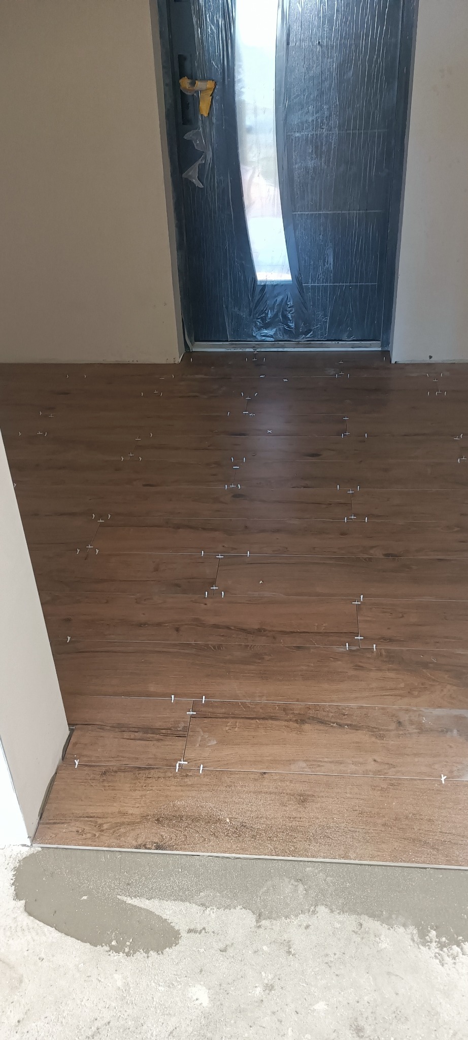 Układanie podłogi z płytek imitujących drewno, z użyciem krzyżyków dystansowych, widoczny próg i zabezpieczone folią drzwi.