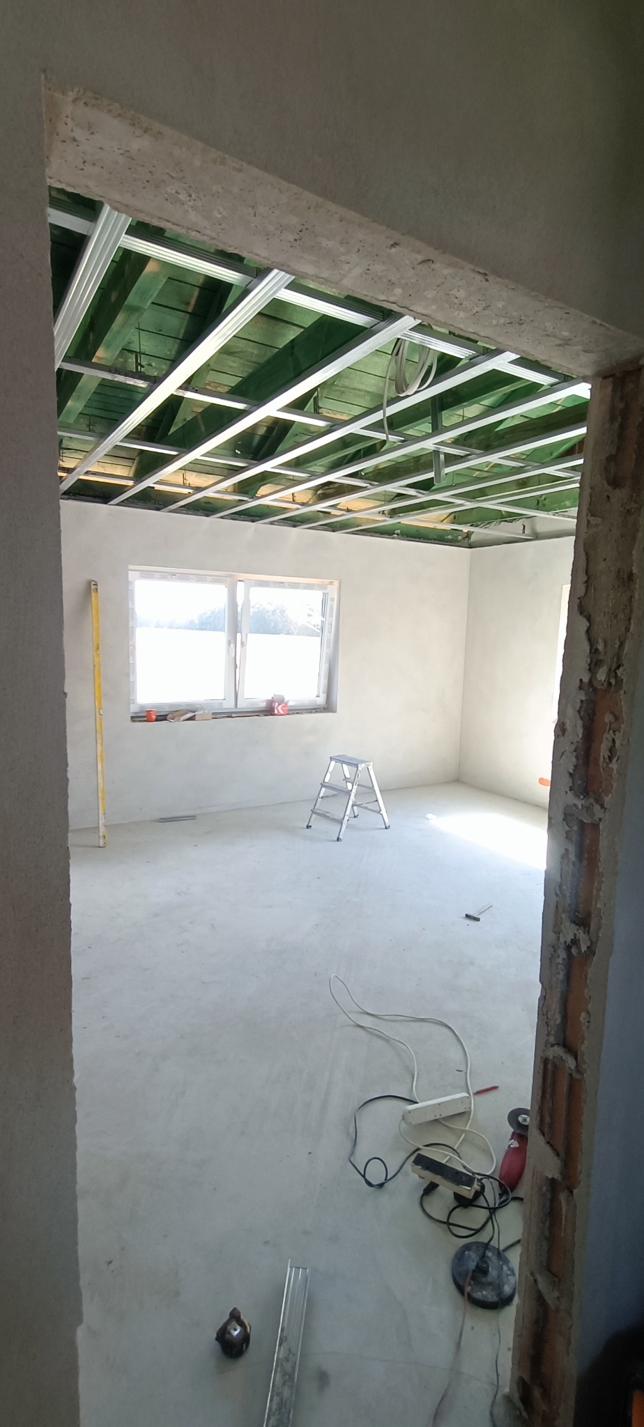 Wnętrze w trakcie remontu: widoczna konstrukcja sufitu podwieszanego z profili metalowych, gładkie ściany przygotowane pod malowanie, okno z widokiem na zewnątrz, drabina aluminiowa i narzędzia...