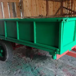 Przyczepa rolnicza po renowacji, świeżo pomalowana na zielono, z czerwonym podwoziem, stojąca w drewnianym budynku gospodarczym.