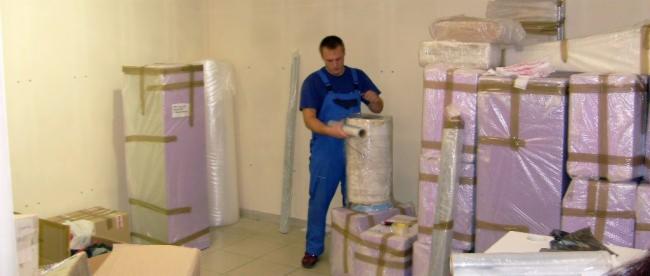 Pracownik w niebieskim kombinezonie zabezpiecza folią stretch okrągły przedmiot przed przeprowadzką, w tle spakowane i oklejone taśmą meble i kartony.