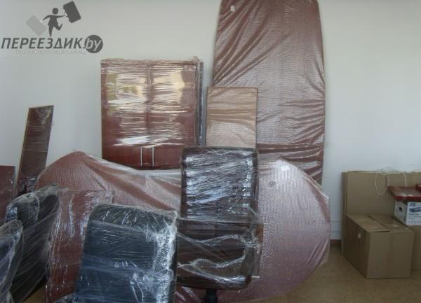 Meble biurowe i domowe, w tym krzesła, stół i szafa, zabezpieczone folią bąbelkową przed transportem, z widocznym logo firmy przeprowadzkowej na ścianie.