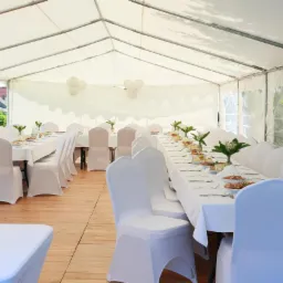 Uroczysty biały namiot bankietowy z drewnianą podłogą, długie stoły nakryte białymi obrusami, białe krzesła w pokrowcach, dekoracje kwiatowe i balony.