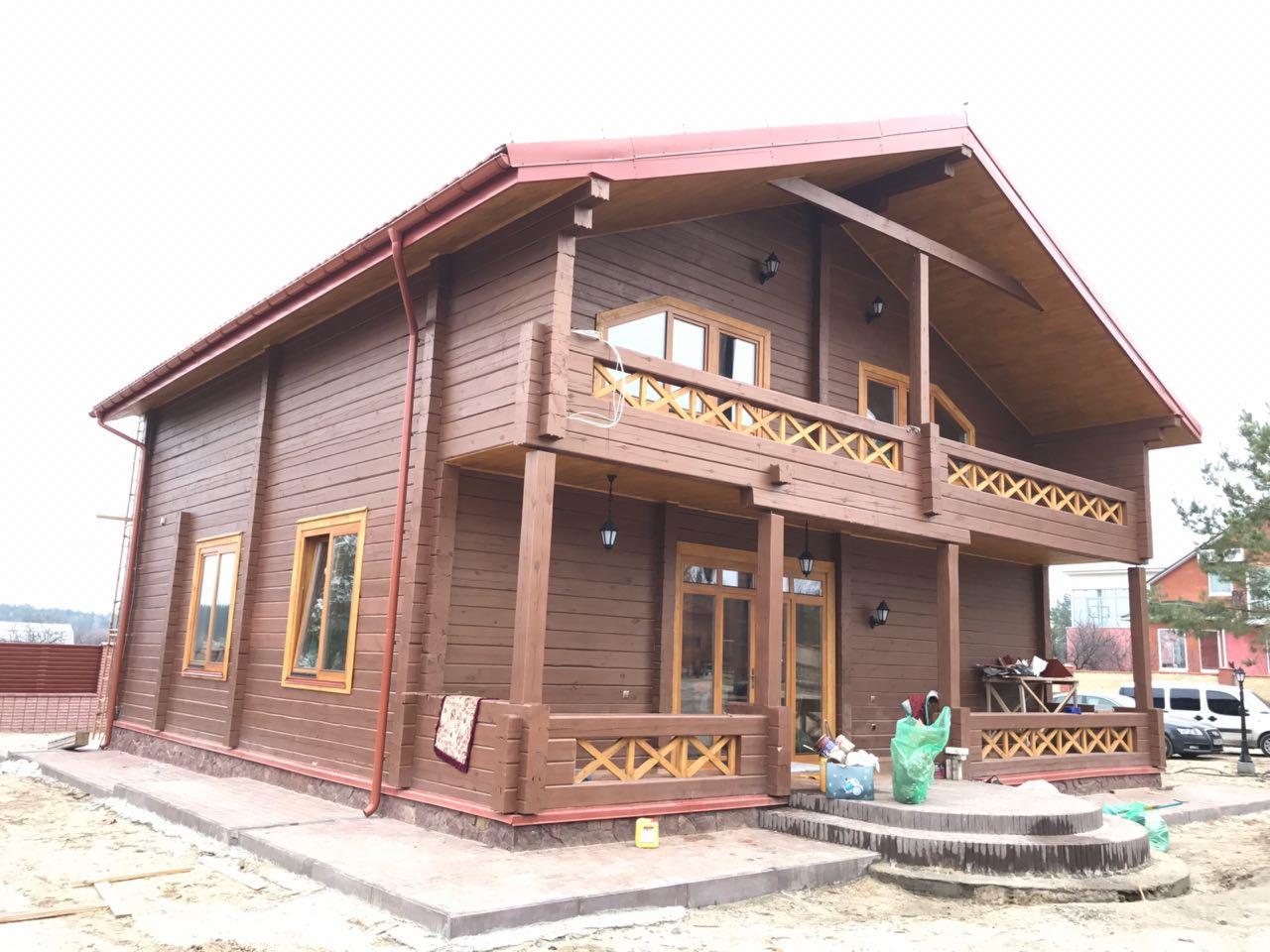 Gotowy, dwukondygnacyjny dom z bali z czerwonym dachem i drewnianą balustradą balkonu, widoczny z zewnątrz, z elementami wykończeniowymi na tarasie.