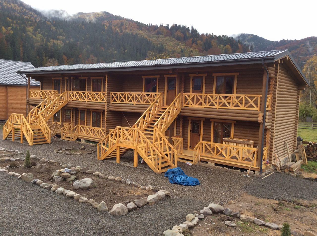 Dwukondygnacyjny budynek z bali z zewnętrznymi schodami z surowego drewna, na tle zalesionych wzgórz i ogrodu z kamiennymi obrzeżami.