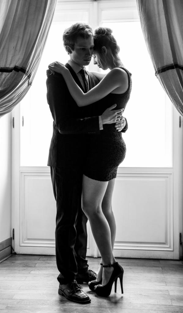 Czarno-białe zdjęcie obejmującej się pary w eleganckich strojach na tle okna z zasłonami.