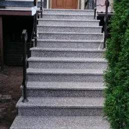 Zewnętrzne schody wejściowe wyłożone szarym, nakrapianym kamieniem z czarną, kutą balustradą, prowadzące do brązowych drzwi wejściowych.