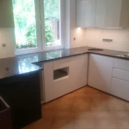 Nowoczesna kuchnia z czarnym, błyszczącym blatem, białymi szafkami i oknem z widokiem na zieleń, brązowa terakota na podłodze.