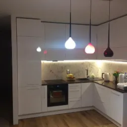 Nowoczesna, biała kuchnia z blatem w kolorze ciemnego brązu, podświetlanym marmurkowym panelem nad blatem i trzema wiszącymi lampami o różnej barwie światła: białej, różowej i czerwonej.