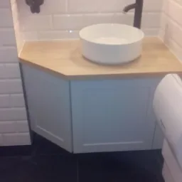 Biała, okrągła umywalka nablatowa z czarną baterią na blacie z jasnego drewna, pod nią szafka w kolorze gołębim, białe kafelki na ścianie, czarna podłoga.