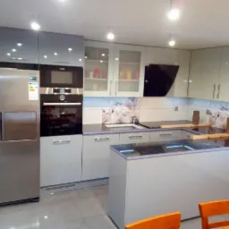 Nowoczesna kuchnia z szarymi, lakierowanymi frontami, wysoka zabudowa z lodówką side-by-side i piekarnikiem w zabudowie, wyspa z czarnym blatem z podświetleniem LED, na blacie roboczym drewniane...