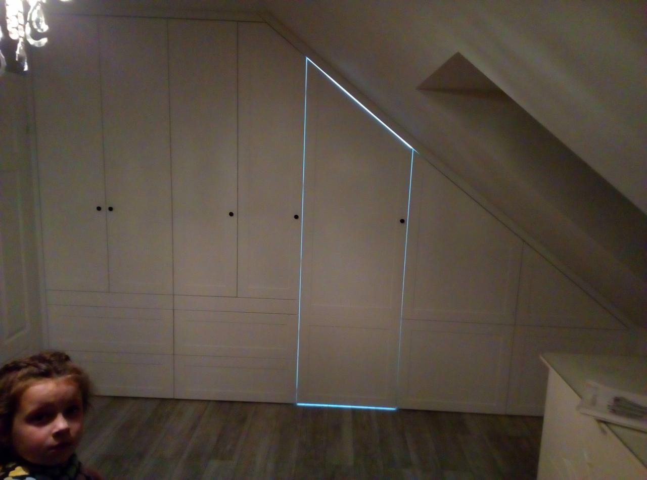 Biała zabudowa wnękowa pod skosem na poddaszu, widoczna twarz dziecka w lewym dolnym rogu, drewniana podłoga.