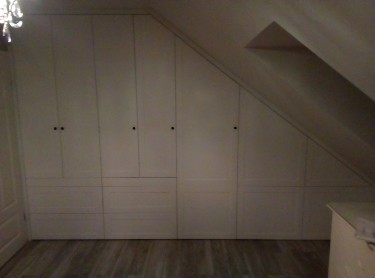 Biała zabudowa szafy wnękowej pod skosem dachu, z licznymi drzwiczkami i szufladami, na podłodze panele imitujące drewno.