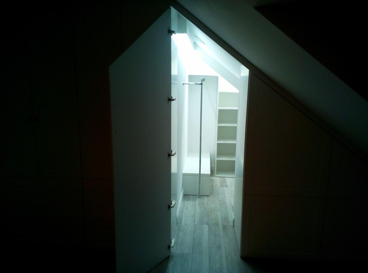 Otwarta biała szafa wnękowa na poddaszu ze skośnym sufitem i półkami, widok z ciemnego pomieszczenia.