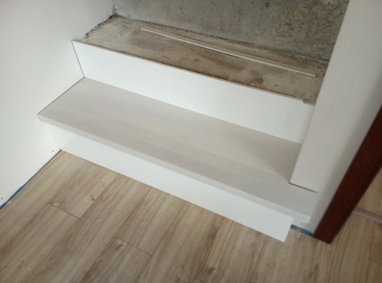 Fragment nowo wykończonego, drewnianego stopnia schodów w jasnej kolorystyce, z widoczną surową ścianą i drewnianą podłogą.