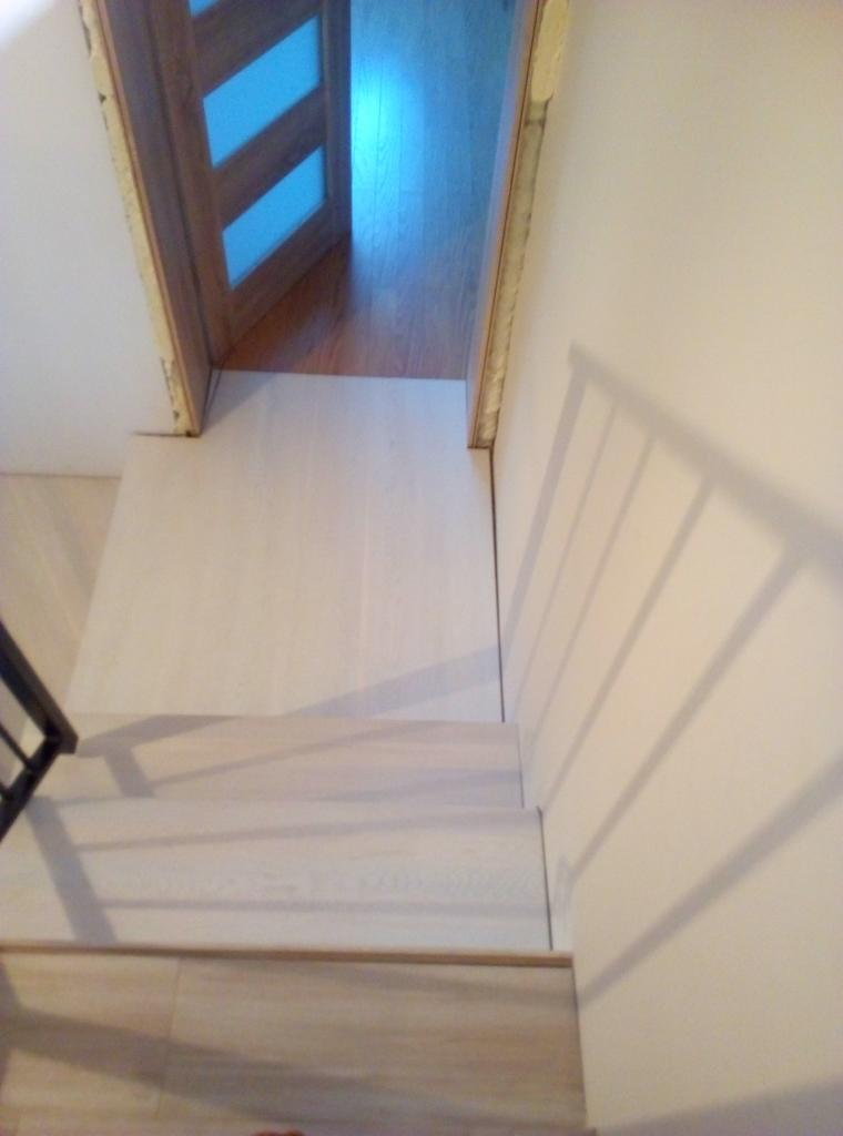 Jasne, drewniane schody prowadzące do drzwi z szybami, widoczne z góry, z cieniem balustrady na ścianie.