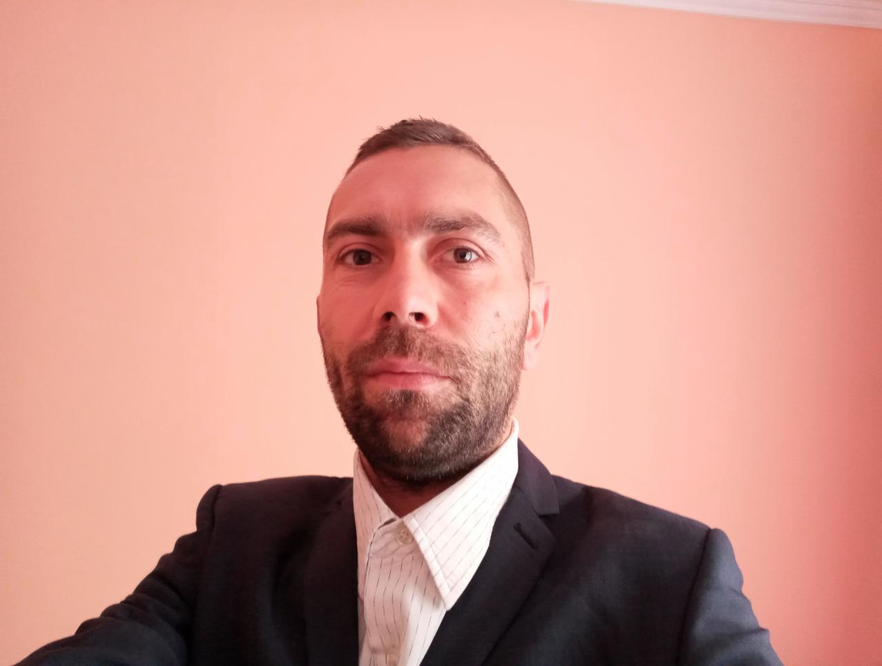 Portret mężczyzny z brodą w garniturze na tle jednolitej, brzoskwiniowej ściany. Mężczyzna patrzy prosto w obiektyw.