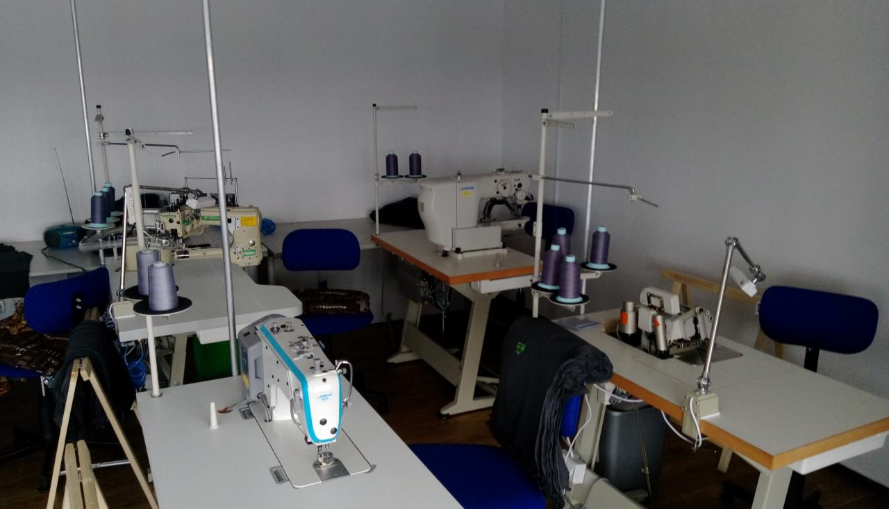 Wnętrze pracowni krawieckiej z kilkoma stanowiskami wyposażonymi w maszyny do szycia, nici w odcieniach fioletu i niebieskie krzesła obrotowe. Na pierwszym planie nowoczesna maszyna z niebieskimi...