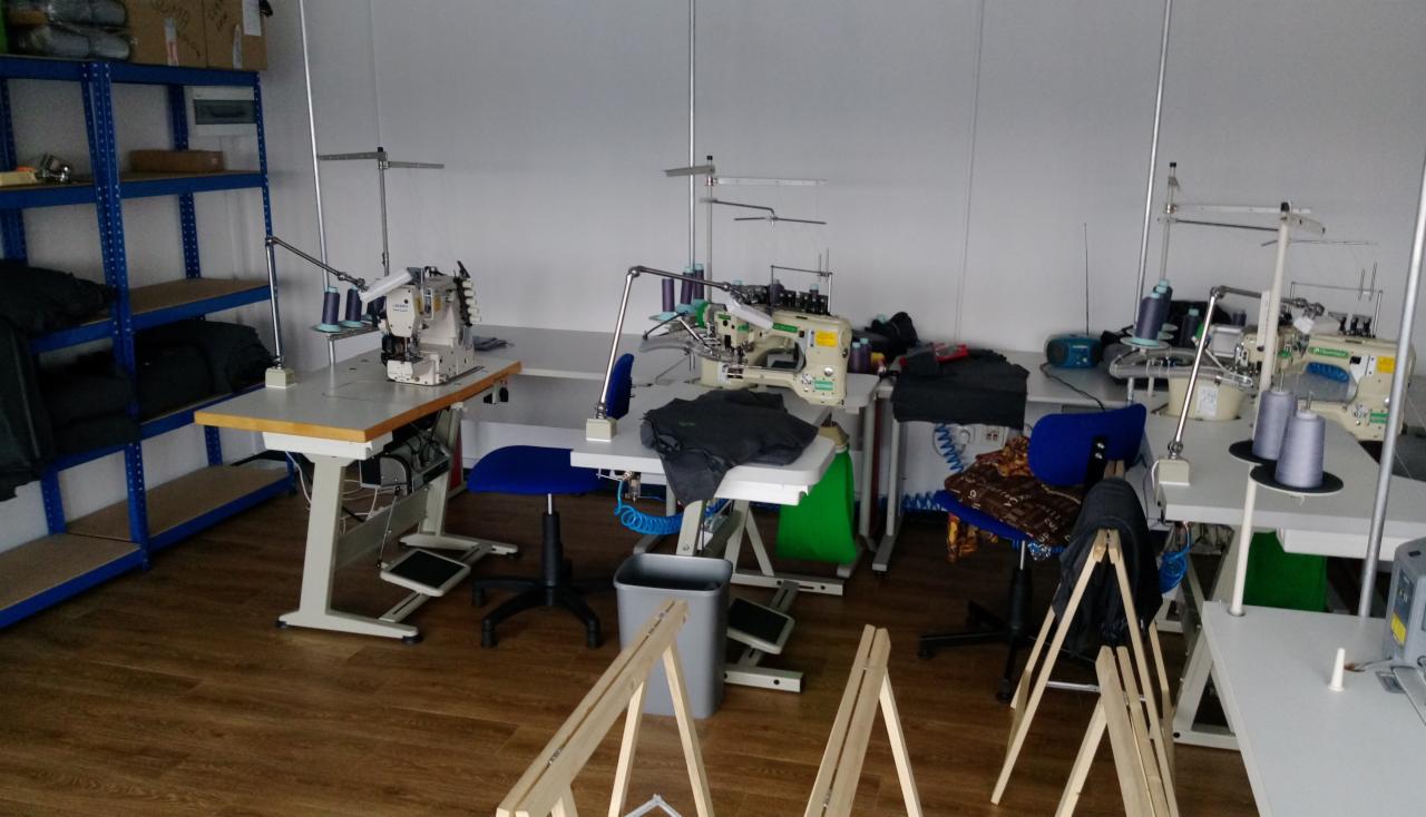 Przestrzeń robocza z trzema stanowiskami krawieckimi wyposażonymi w maszyny do szycia przemysłowego, krzesła obrotowe, metalowe regały z gotowymi produktami oraz drewniane kozły malarskie...