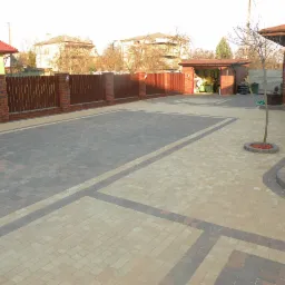 Ułożona kostka brukowa w dwóch odcieniach szarości i beżu tworząca wzór na podjeździe, z widocznym ogrodzeniem z cegły i drewna oraz garażem w tle.